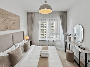 Une chambre moderne avec un grand lit, une table de chevet, un miroir et une vue sur la ville à travers des rideaux transparents.