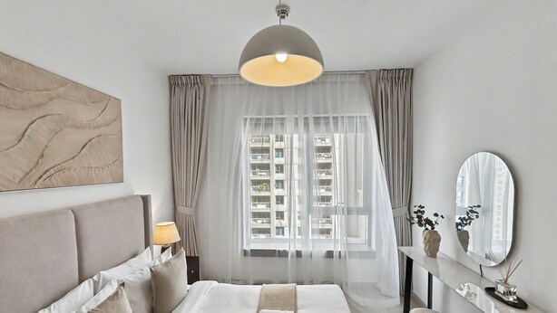 Een moderne hotelkamer met een groot bed, nachtkastje, spiegel en uitzicht op de stad door dunne gordijnen.