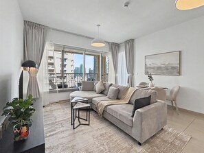 A modern living room with a grey sofa, a small side table, a potted plant, and a view of the city through large windows. - 29 Boulevard T1 - 1801 1BD (Dubai)