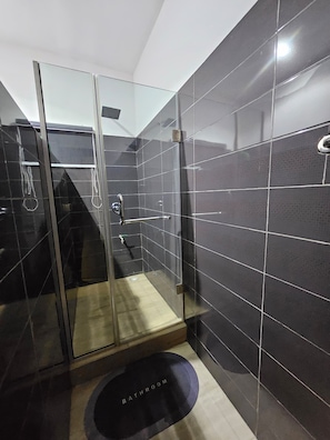 Un baño moderno con paredes de baldosas oscuras, cabina de ducha con mampara de cristal y un dispensador de papel higiénico visible.