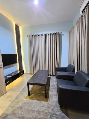 Una sala de estar moderna con un televisor de pantalla plana, una mesa de centro de madera oscura, un sofá gris y un ventanal con cortinas.