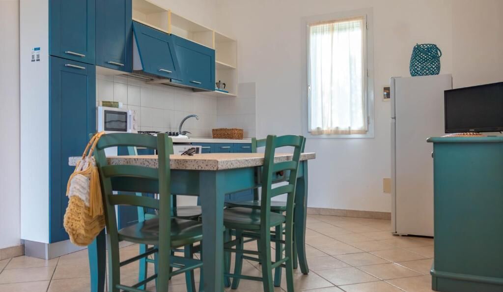 Uma cozinha compacta com armários cor de verde-água, uma mesa de jantar com cadeiras, uma janela com cortinas e uma televisão em um móvel.