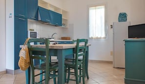 A compact kitchen with teal cabinets, a dining table with chairs, a window with curtains, and a television on a stand.