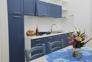 A kitchen with blue cabinets, a gas stove, a sink, and a table with chairs. There is a basket of fruit and a bottle on the counter.