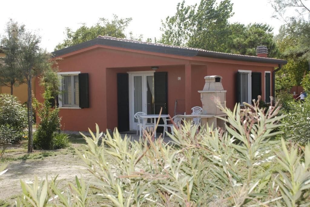 A small building with a red exterior, a door, and a window. There is a white outdoor table and chairs, and a small garden area with plants.