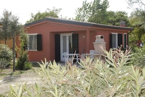 A small building with a red exterior, a door, and a window. There is a white outdoor table and chairs, and a small garden area with plants.