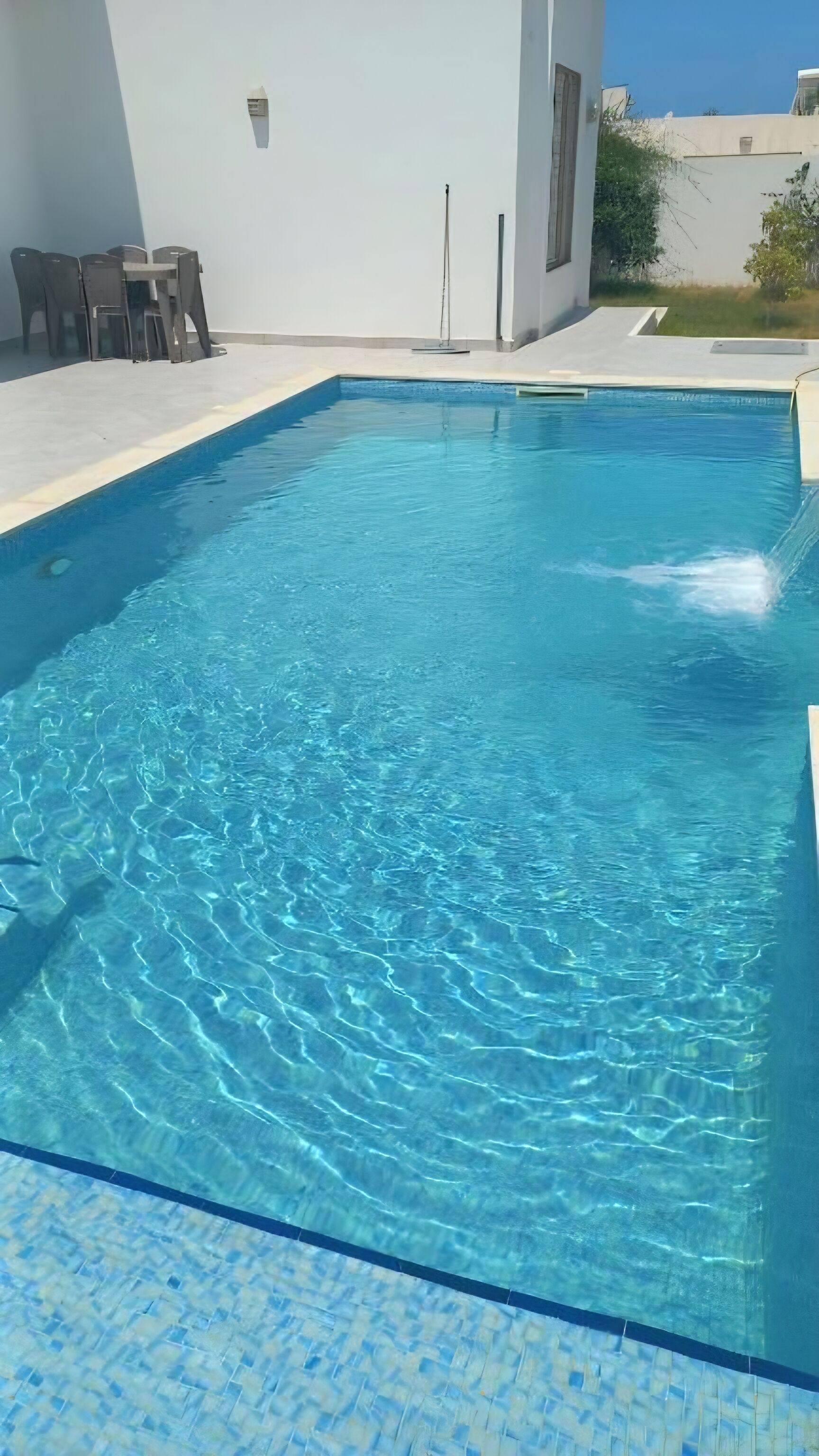 A rectangular swimming pool with clear blue water, surrounded by a tiled deck and a white building with a window.