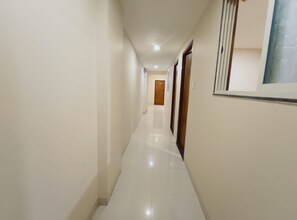 Un pasillo con una puerta de madera al fondo, paredes blancas y suelo de baldosas.