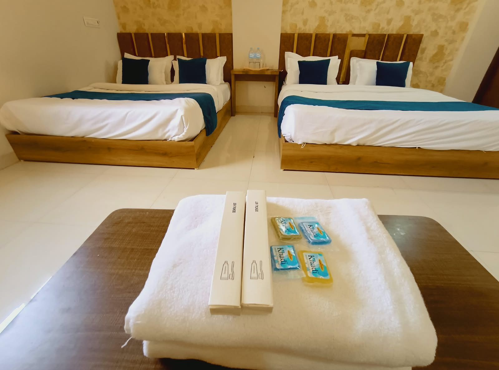 Habitació d'hotel amb dos llits, un panell de capçalera de fusta, una tauleta de nit amb una ampolla d'aigua i una tovallola amb articles de tocador.