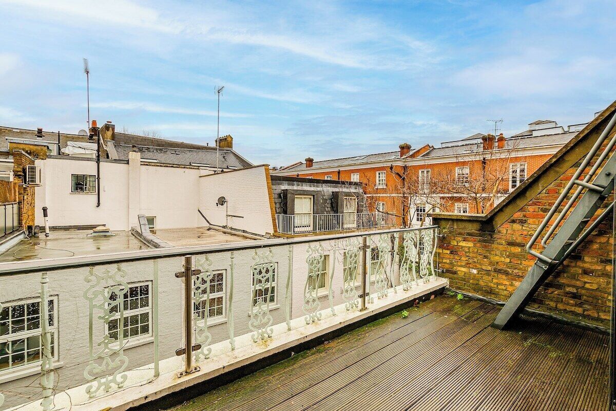A rooftop terrace with a white railing, a view of adjacent buildings, and a clear sky.