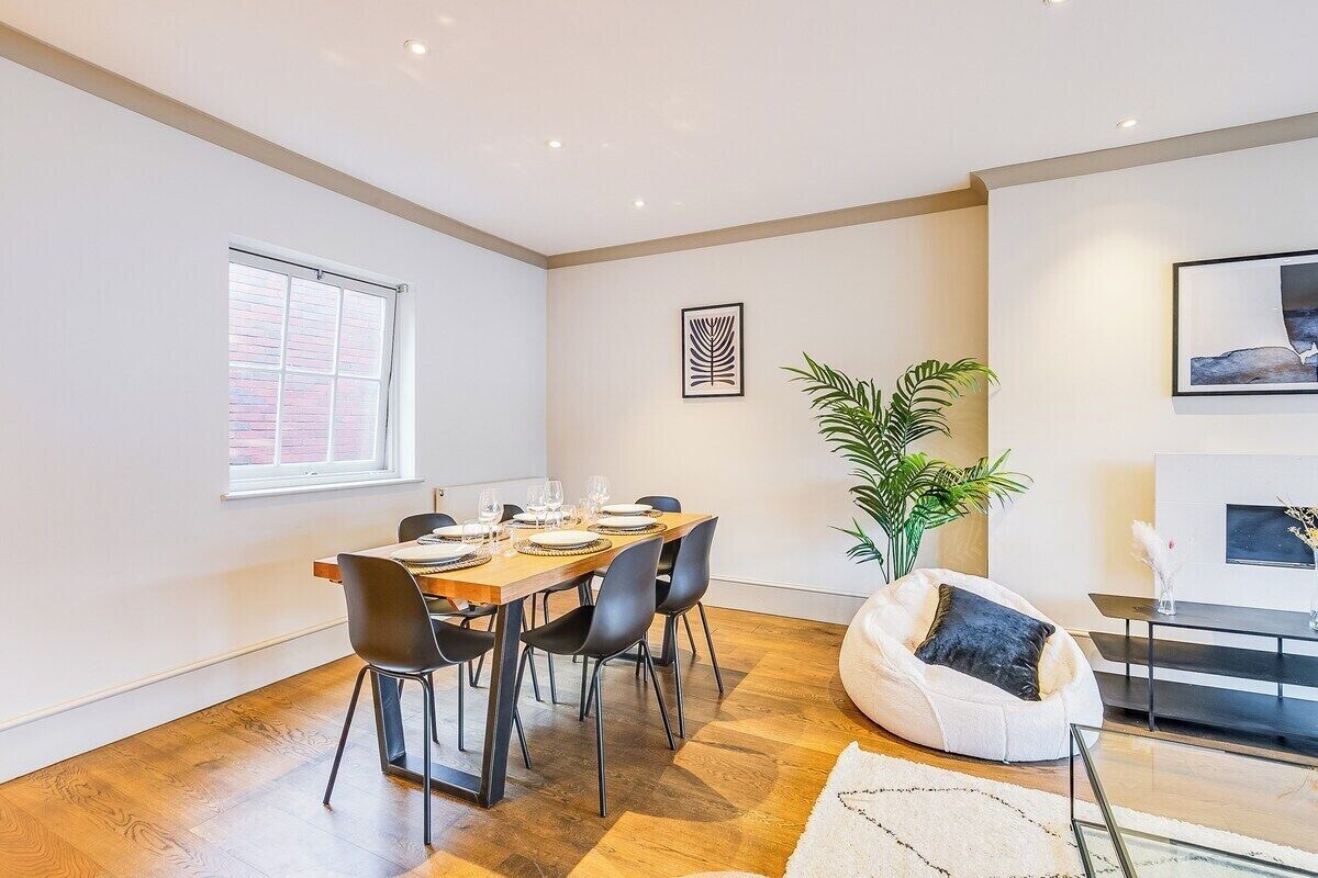 Un comedor moderno con una mesa de madera dispuesta para cuatro, sillas negras, una planta en maceta y un sillón de cuero tipo bean bag.
