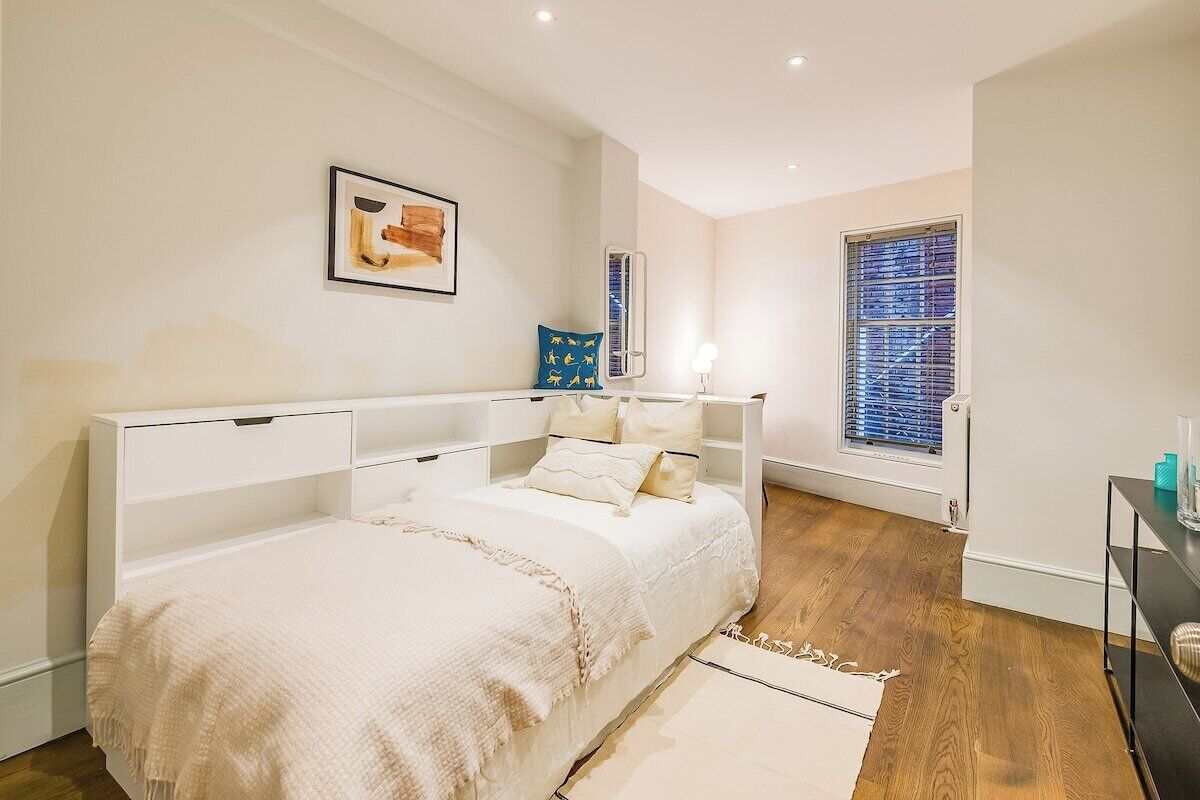 Une chambre à coucher comprenant un lit, des tables de chevet, une fenêtre avec des stores et une œuvre d’art accrochée au mur.