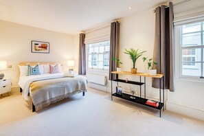 Une chambre d’hôtel comprenant un lit, des tables de chevet, une lampe de chevet, une fenêtre avec des rideaux, une étagère fixée au mur avec des livres et une plante en pot.