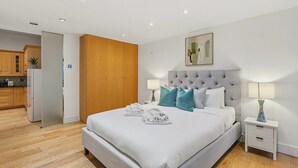 Un dormitorio con una cama grande, una mesita de noche, un armario de madera y una cocina al fondo.