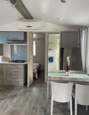 A compact living space with a kitchenette, a dining area with two chairs, and a bedroom with a bed. - Cozy-house 2-bedroom - Les Embruns 4* Campground (Lège-Cap-Ferret)