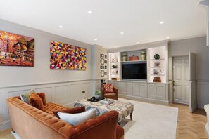 Ein Wohnzimmer mit braunem Sofa, einem Couchtisch und einem Fernseher auf einem Schrank. An den Wänden hängen zwei Bilder und ein Bücherregal mit Dekorationen.