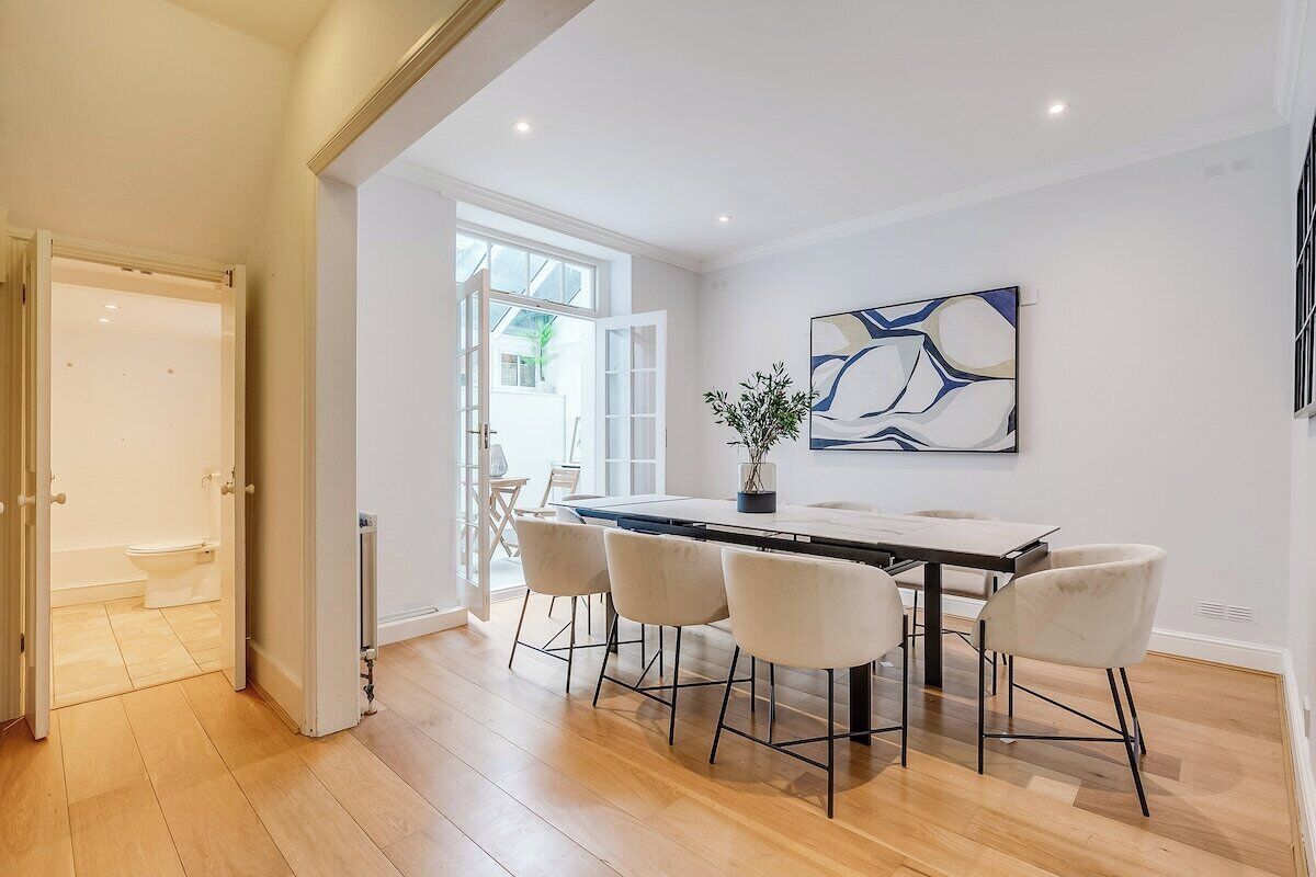 A modern dining area with a long table and four chairs, a potted plant, and a contemporary abstract painting on the wall.