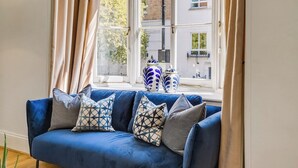 Un canapé bleu avec des coussins à motifs, une fenêtre avec des rideaux et une vue sur les bâtiments à l’extérieur.
