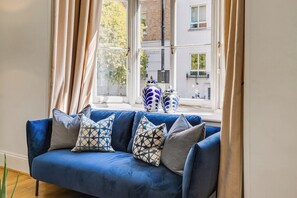 Ein blaues Sofa mit gemusterten Kissen, ein Fenster mit Vorhängen und ein Blick auf Gebäude im Freien.