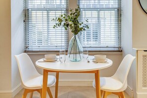 A dining area with a round table, two white chairs, a vase with green foliage, and a window with blinds. - Belgravia - Charming   Spacious 4 Bed Home for 9 (Greater London)