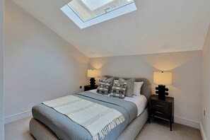 Ein Schlafzimmer mit Dachfenster, einem Bett mit grau-weißer Bettdecke, zwei Nachttischen mit Lampen und einer gemusterten Kissenanordnung.