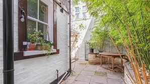 Un patio estrecho con un edificio blanco, una mesa de madera y plantas de bambú.