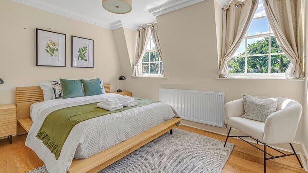 Un dormitorio con cama, mesitas de noche, una silla, una ventana con cortinas y cuadros en la pared.
