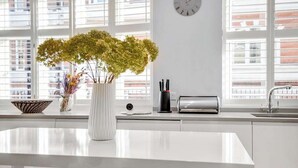 Uma cozinha moderna com bancada branca, um vaso com flores, um relógio e uma janela com persianas.