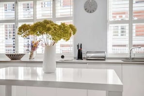 Eine moderne Küche mit weißer Arbeitsplatte, einer Blumenvase, einer Uhr und einem Fenster mit Jalousien.