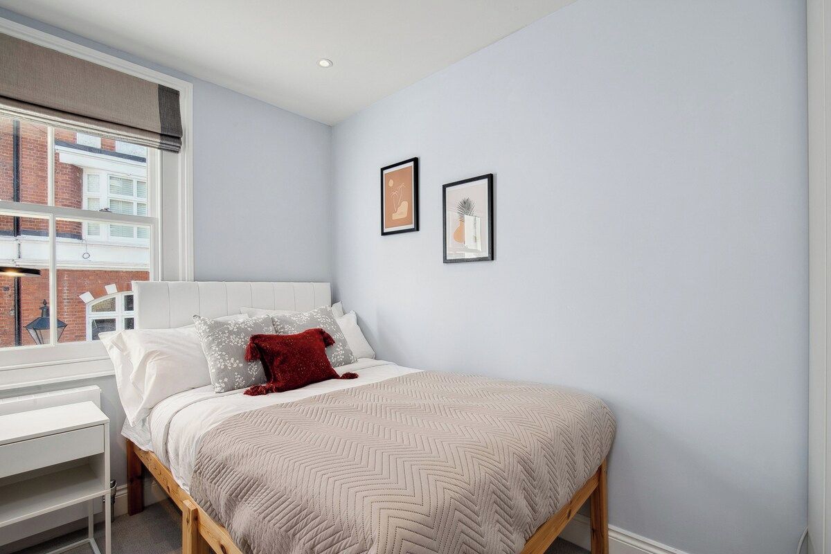 A bedroom with a bed, a window, a desk, and two framed pictures on the wall.