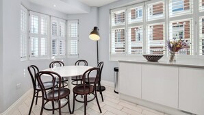 Uma sala de jantar com uma mesa e cadeiras brancas, um abajur e uma janela com persianas dando para um prédio de tijolos.