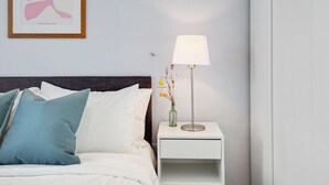 Uma cama bem arrumada com almofadas, uma mesa de cabeceira com abajur e uma obra de arte emoldurada na parede.