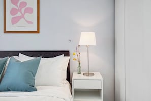 Ein ordentlich gemachtes Bett mit Kissen, ein Nachttisch mit einer Lampe und ein gerahmtes Kunstwerk an der Wand.