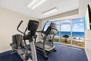 Ein Fitnessraum mit Laufbändern und einem Ergometer, große Fenster mit Blick auf den Strand und ein blauer Teppich.
