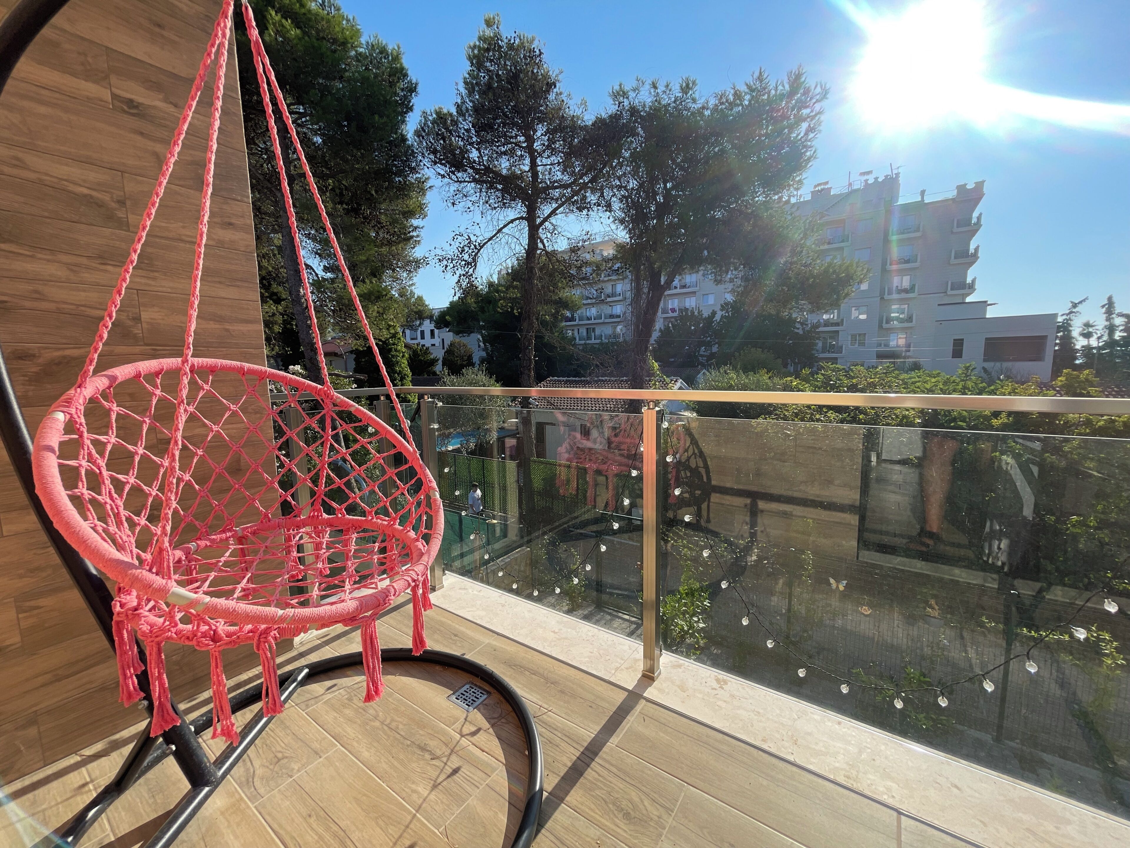 A red macrame chair hanging from a balcony railing, with a view of a building and trees in the background.