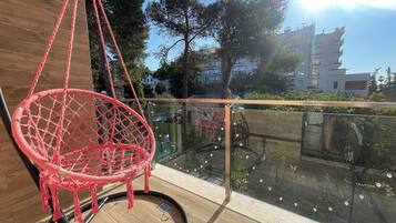 A red macrame chair hanging from a balcony railing, with a view of a building and trees in the background.