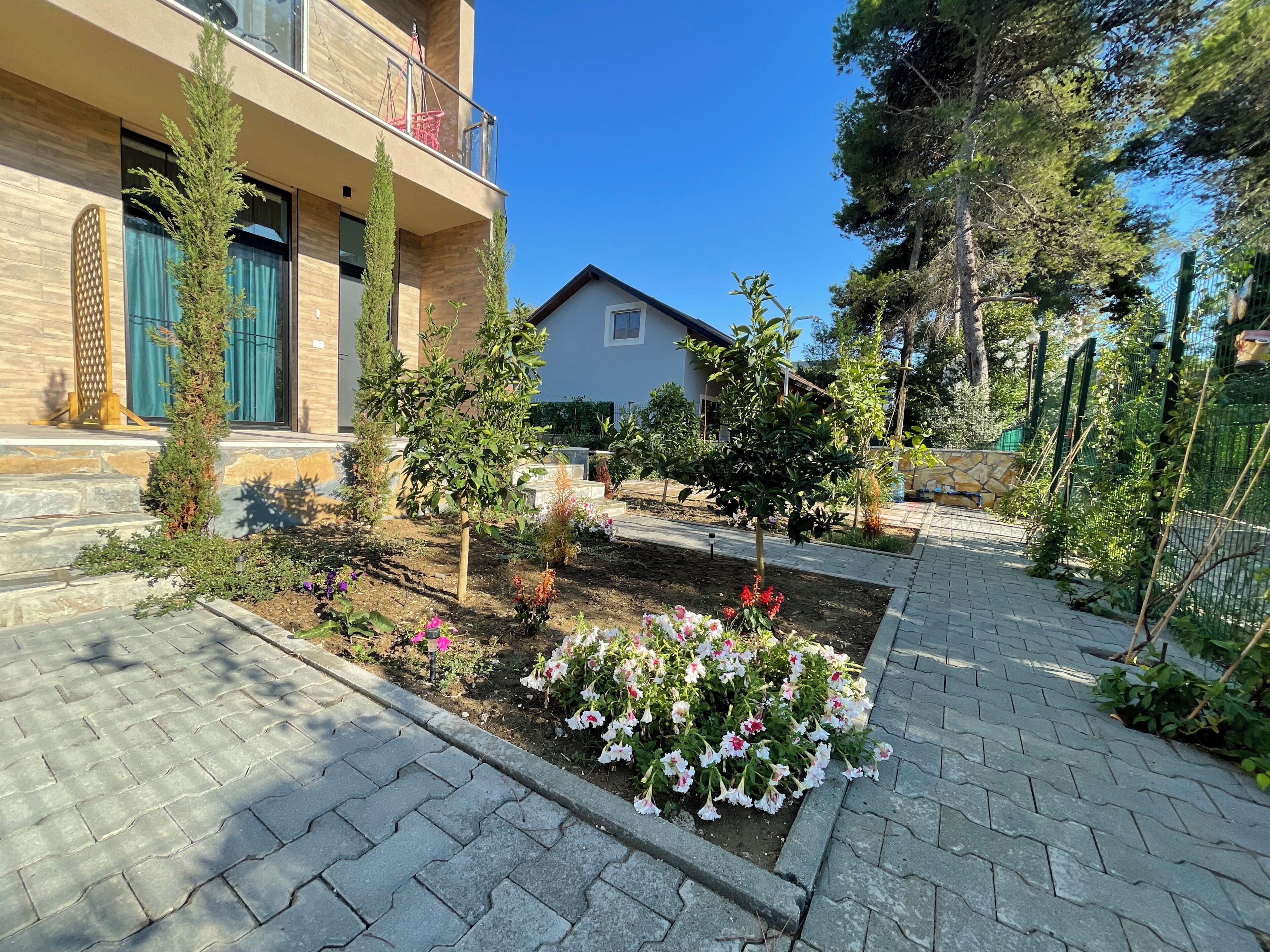 Um bairro residencial com um caminho pavimentado, um jardim com diversas plantas e flores, e um prédio com varanda.