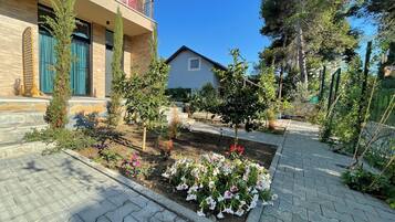 Um bairro residencial com um caminho pavimentado, um jardim com diversas plantas e flores, e um prédio com varanda.