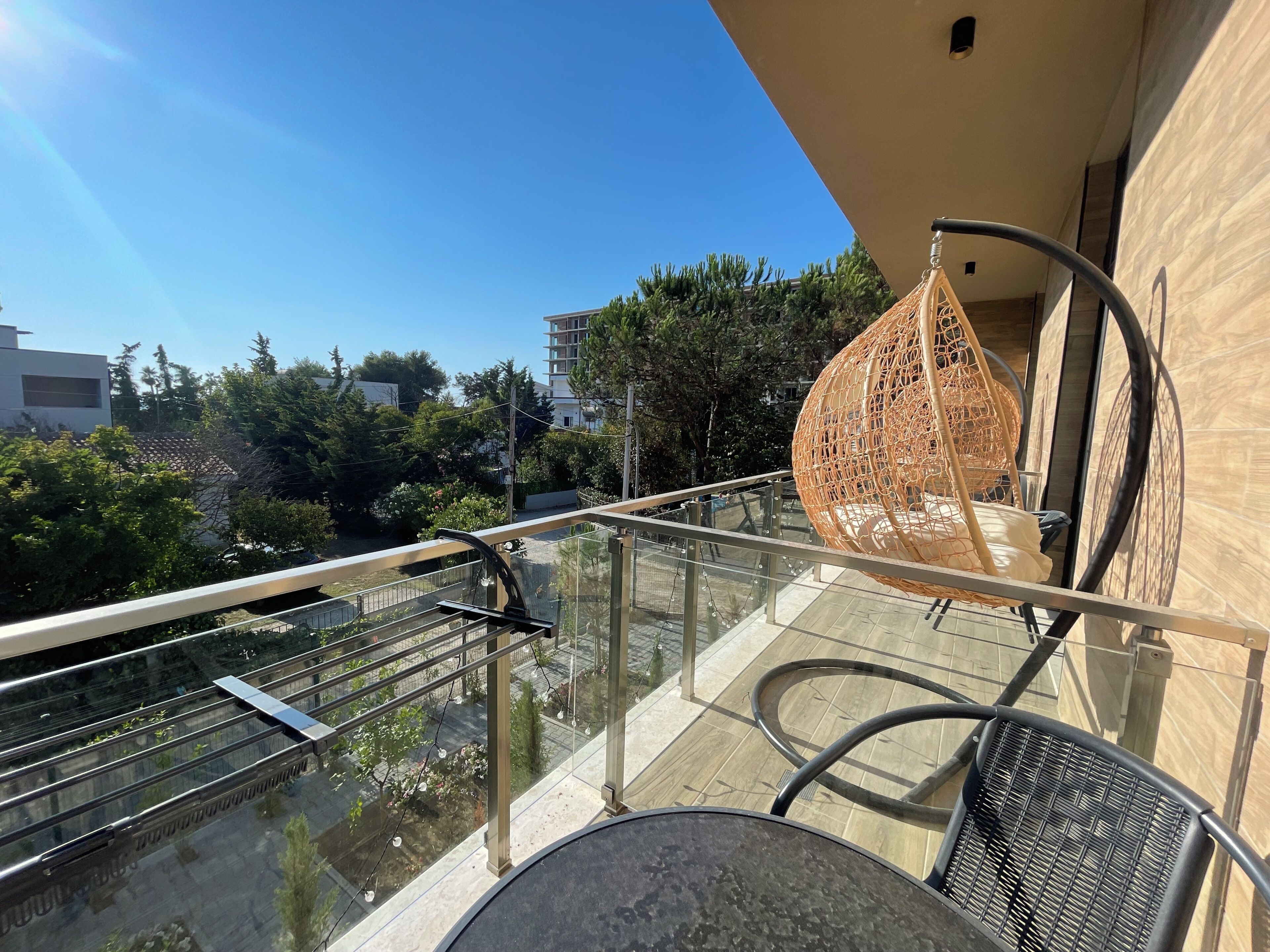 Een balkon met een hangstoel, een tafel en een glazen balustrade met uitzicht op een tuin en gebouwen.