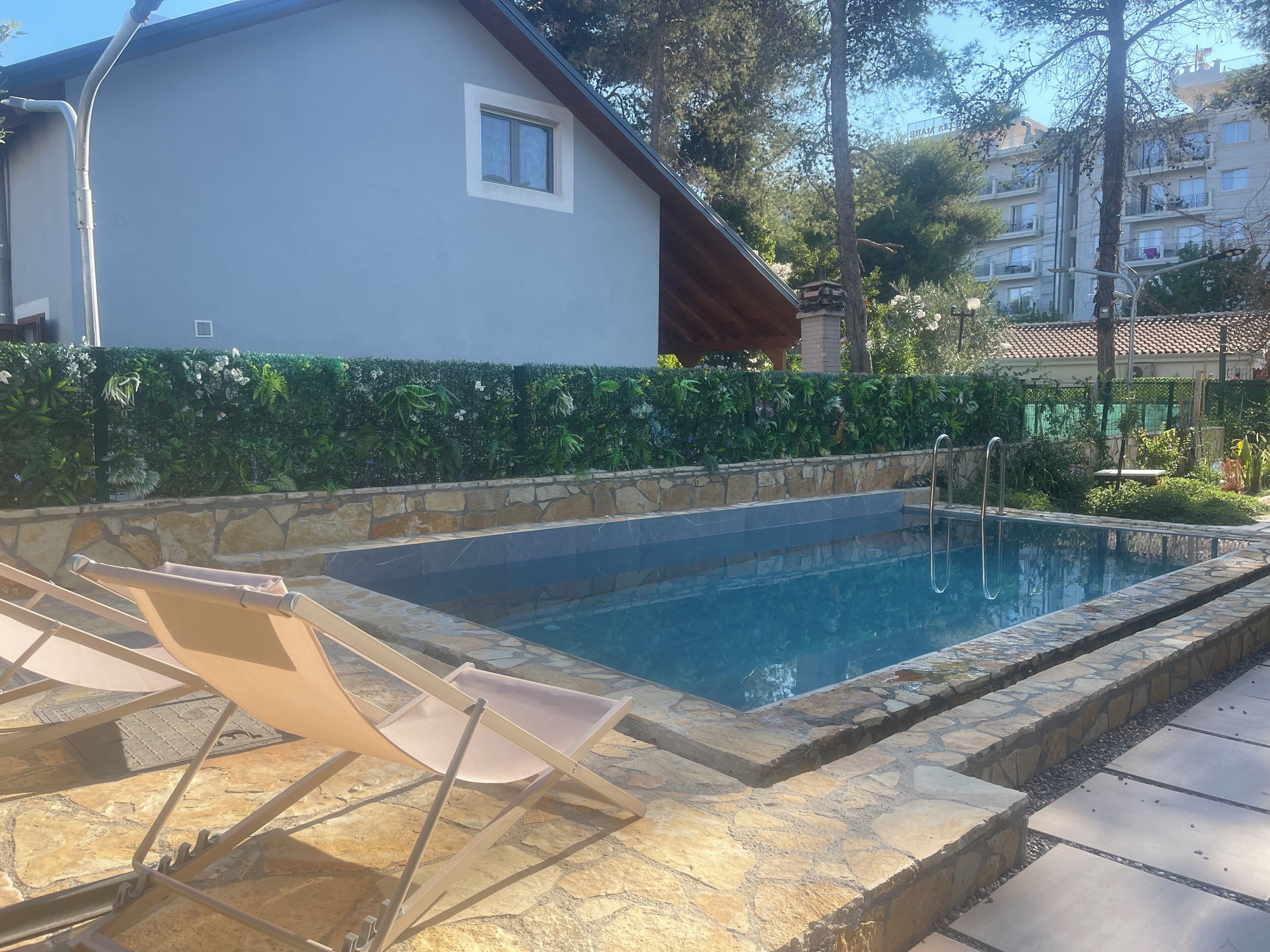 Uma piscina com espreguiçadeiras, um muro de pedra e um edifício ao fundo.