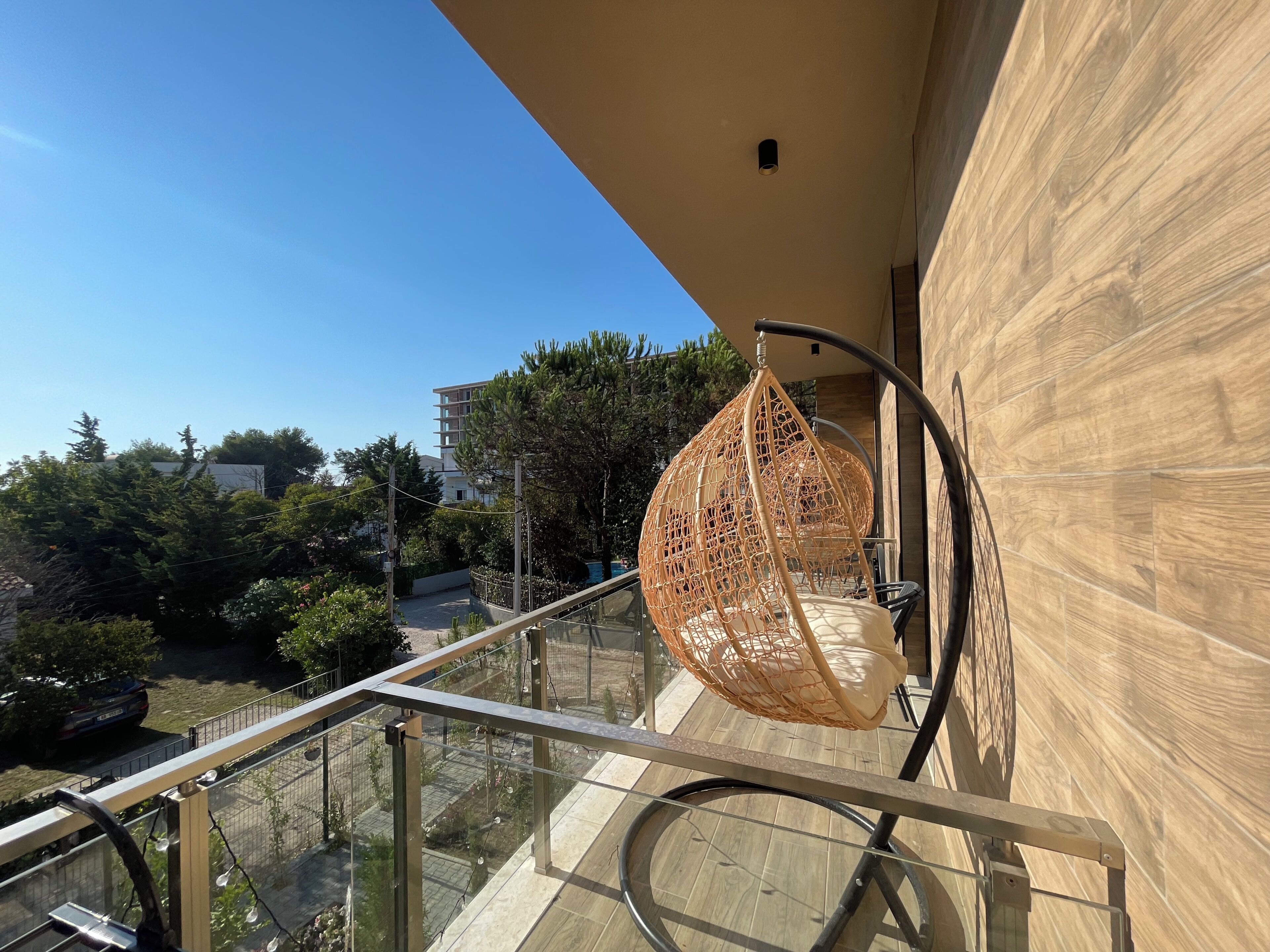 Un balcon doté d’un fauteuil suspendu, d’une rambarde en verre et offrant une vue sur les environs, notamment sur les arbres et les bâtiments.