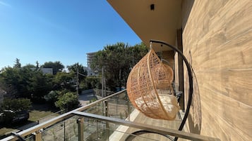 Un balcon doté d’un fauteuil suspendu, d’une rambarde en verre et offrant une vue sur les environs, notamment sur les arbres et les bâtiments.