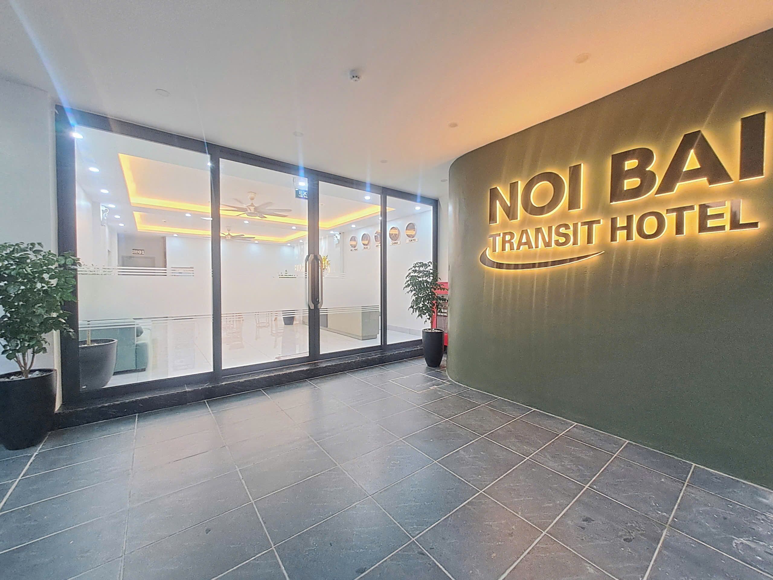 A hotel reception area with a glass door entrance, a reception desk, and a wall with the hotel name illuminated.