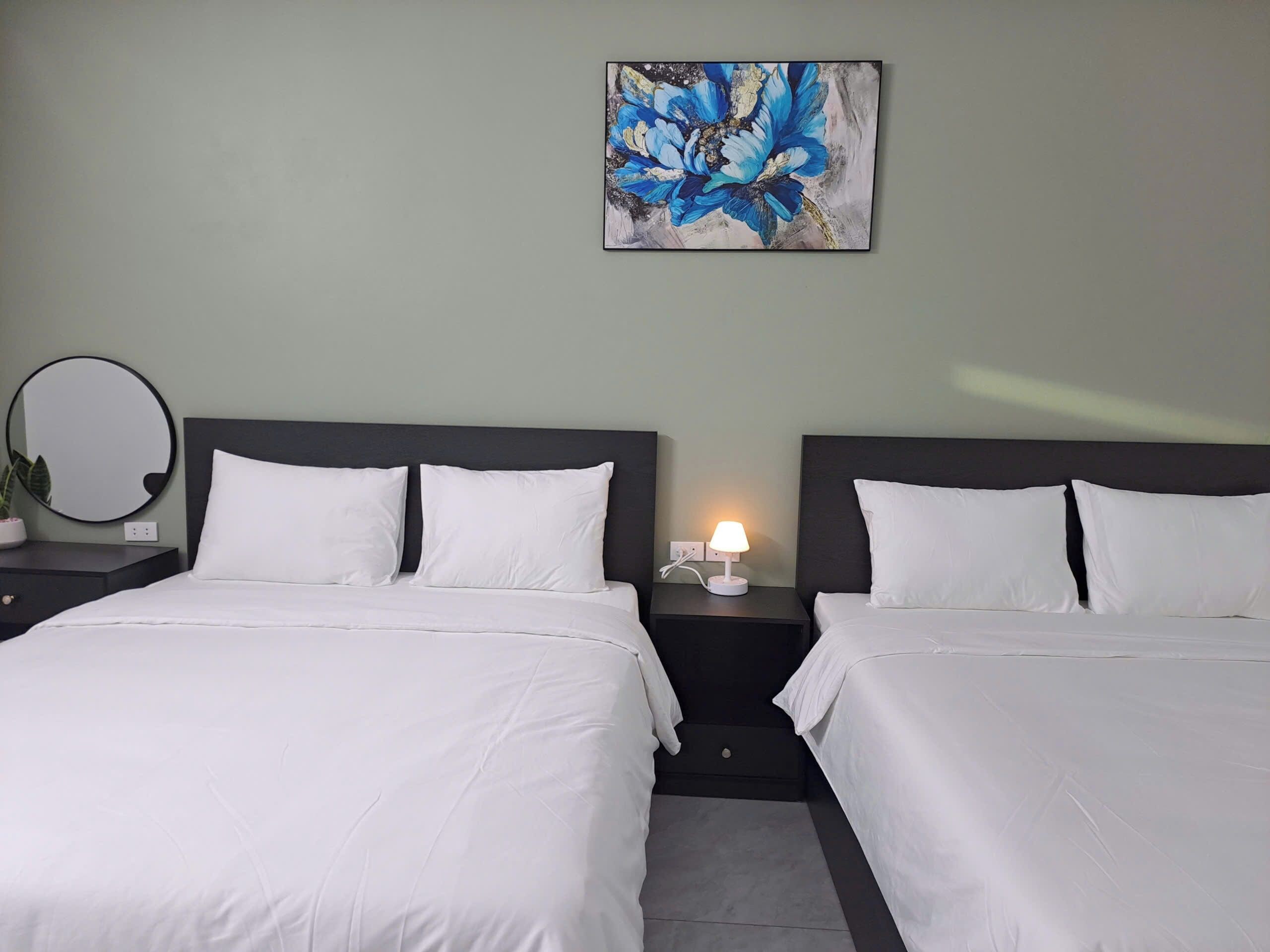 A hotel room with two beds, a bedside table with a lamp, a round mirror, and a painting on the wall.