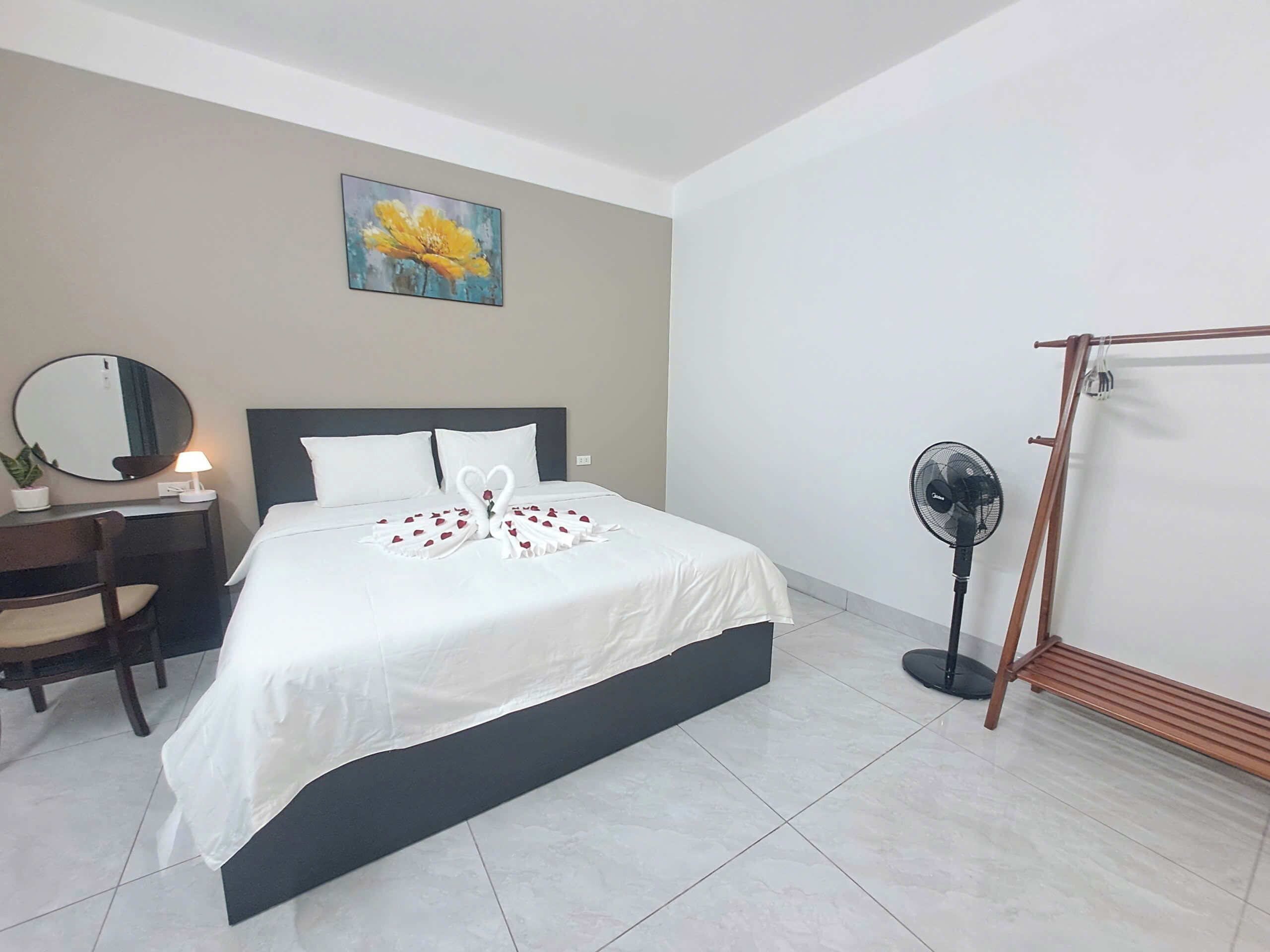 A hotel room with a bed, a desk with a chair, a mirror, a wall-mounted fan, and a painting.