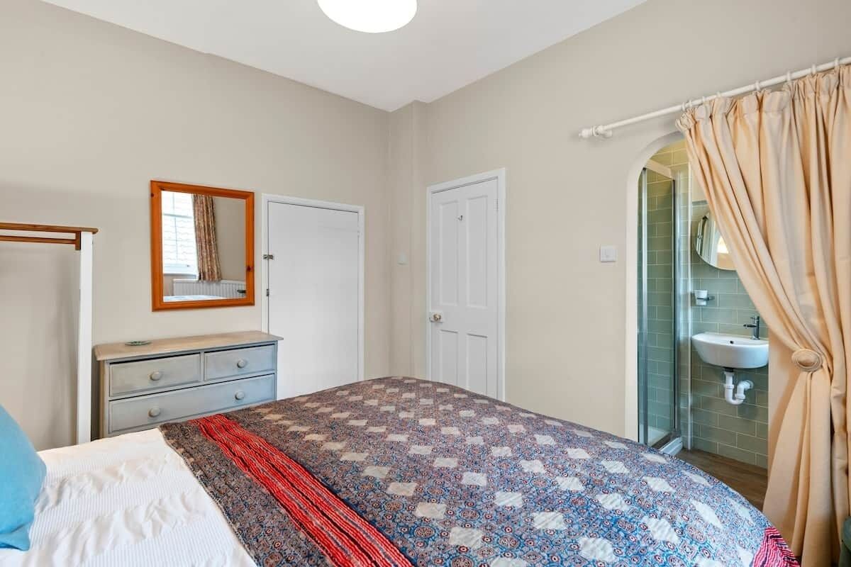 A bedroom with a bed, a dresser, a mirror, a door, and a bathroom with a sink and shower.