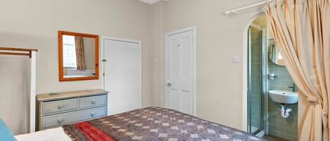 A bedroom with a bed, a dresser, a mirror, a door, and a bathroom with a sink and shower.