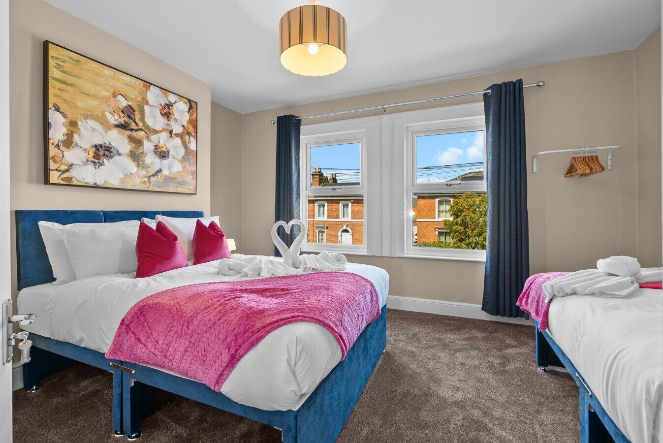 Une chambre d’hôtel avec deux lits, une grande fenêtre avec des rideaux, un tableau au mur et un luminaire au plafond.