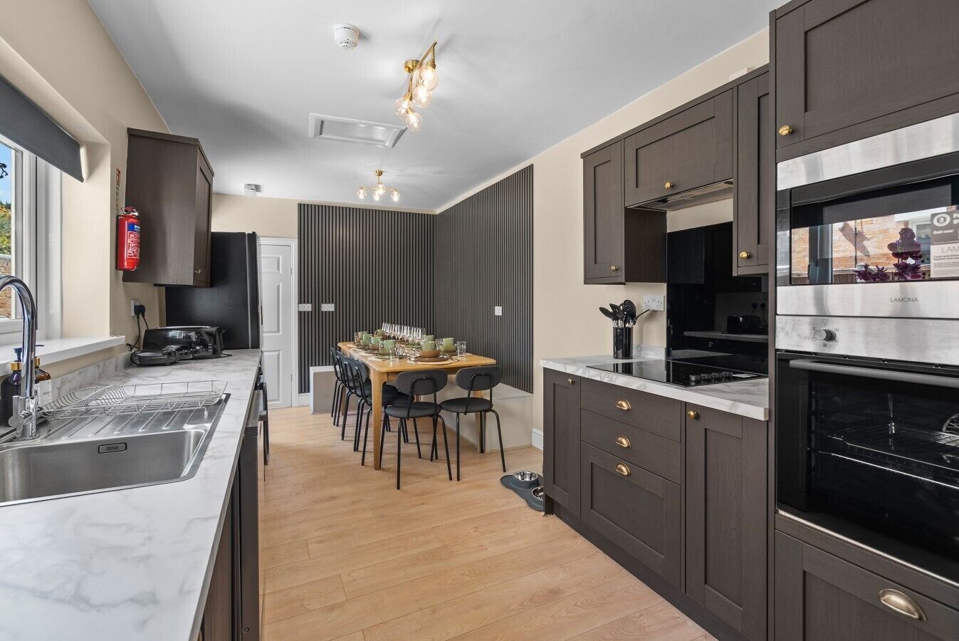 Une cuisine moderne avec des meubles de couleur sombre, un plan de travail en marbre, un four encastré et un coin repas avec une table dressée pour quatre personnes.