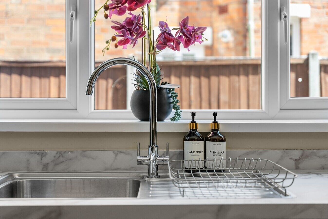 Un évier de cuisine avec un robinet moderne, une plante en pot aux fleurs violettes et deux bouteilles de savon pour les mains et la vaisselle posées sur le comptoir.